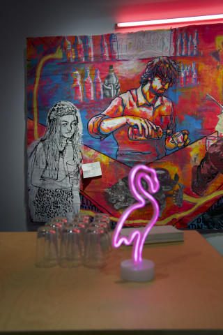 Bar aus Holz mit einem leuchtenen Flamingo und Gläsern. Im Hintergrund eine großformatige Malerei mit einer Ausschenksituation in einer Bar.