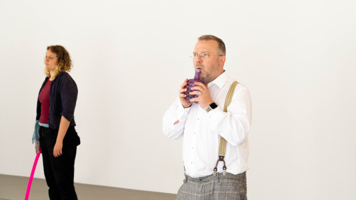 Zwei Personen stehen im leeren Raum. Eine Person pfeift mit Hilfe eines Flaschen ähnlichem Objektes.