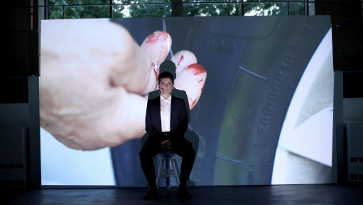 Eine Frau im Anzug sitzt auf einem Hocker und wird von einem Beamer bespielt. Zu sehen sind blutige Finger an einem Reifen