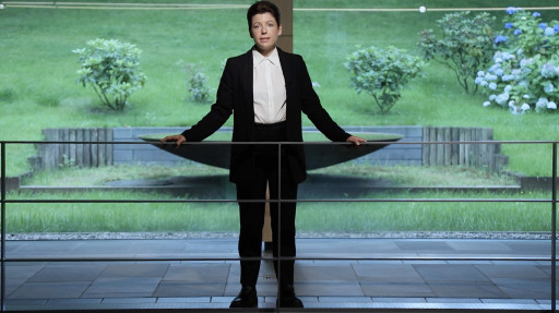 Eine Frau im Anzug steht an einem Geländer mit einem Garten im Hintergrund